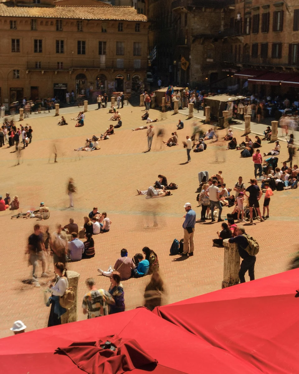 People sitting and walking across the sunlit Piazza del Campo, framed by red awnings.