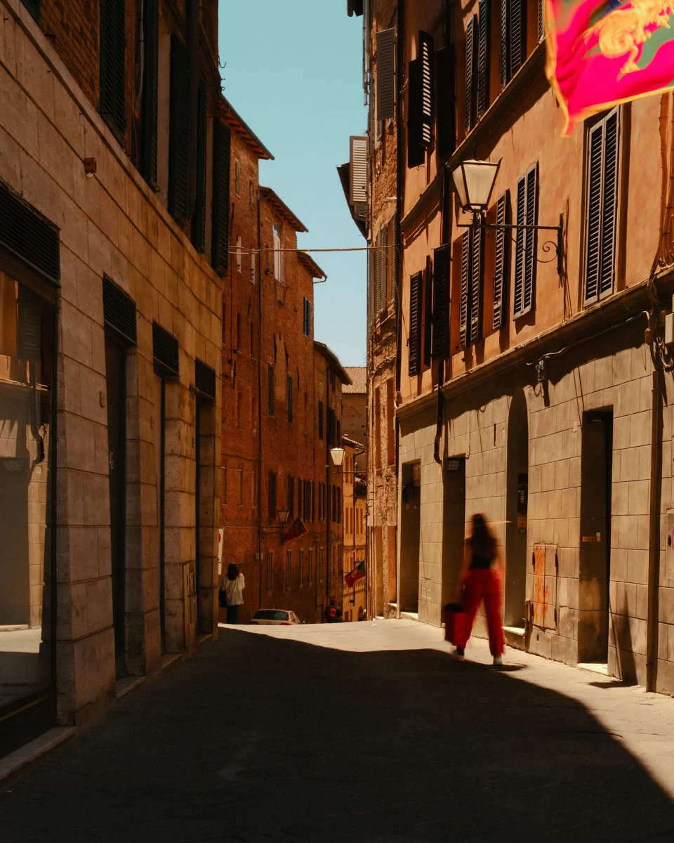 A lone pedestrian on a narrow sunlit street, with a colorful flag overhead.