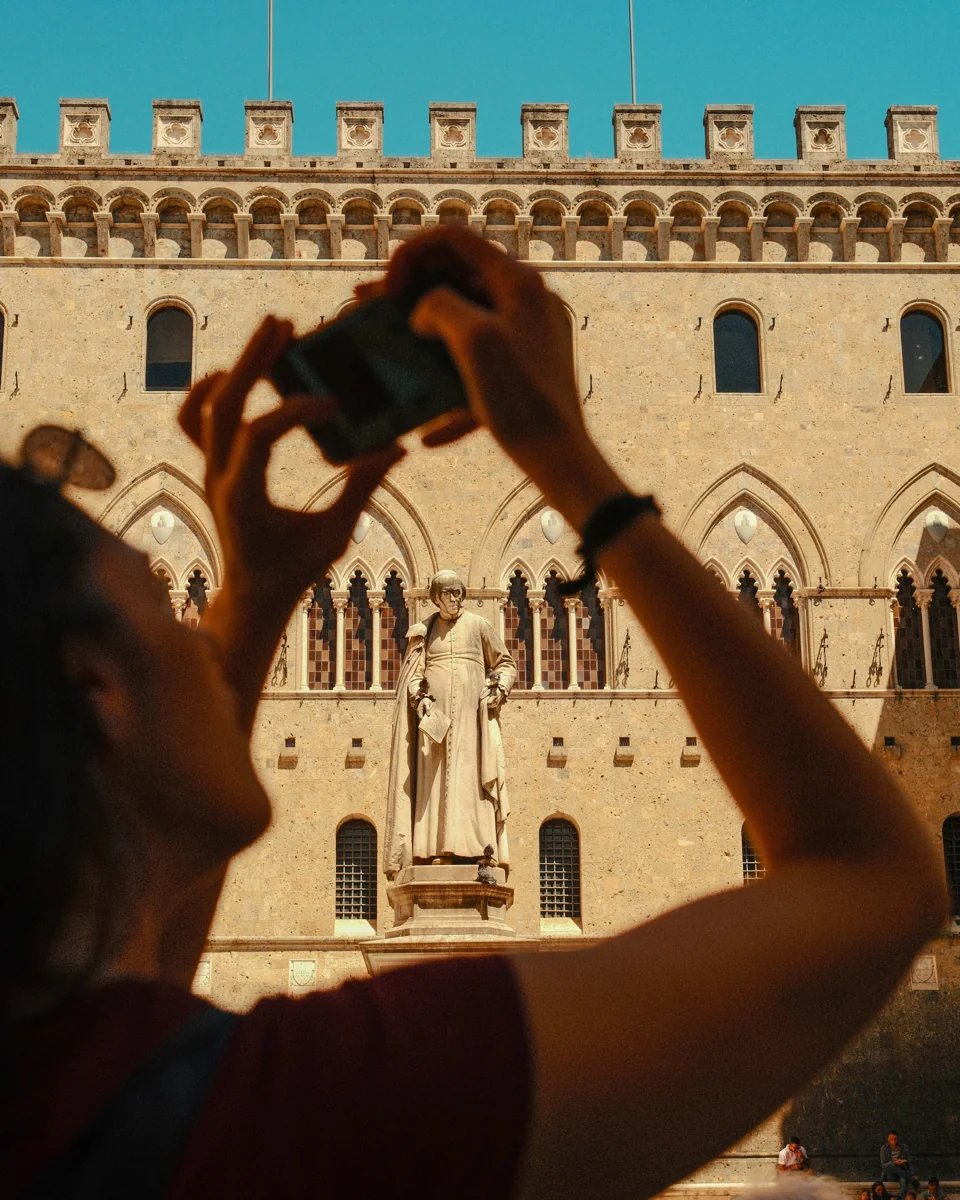 A visitor raising a camera toward an ornate stone building beneath a blue sky.