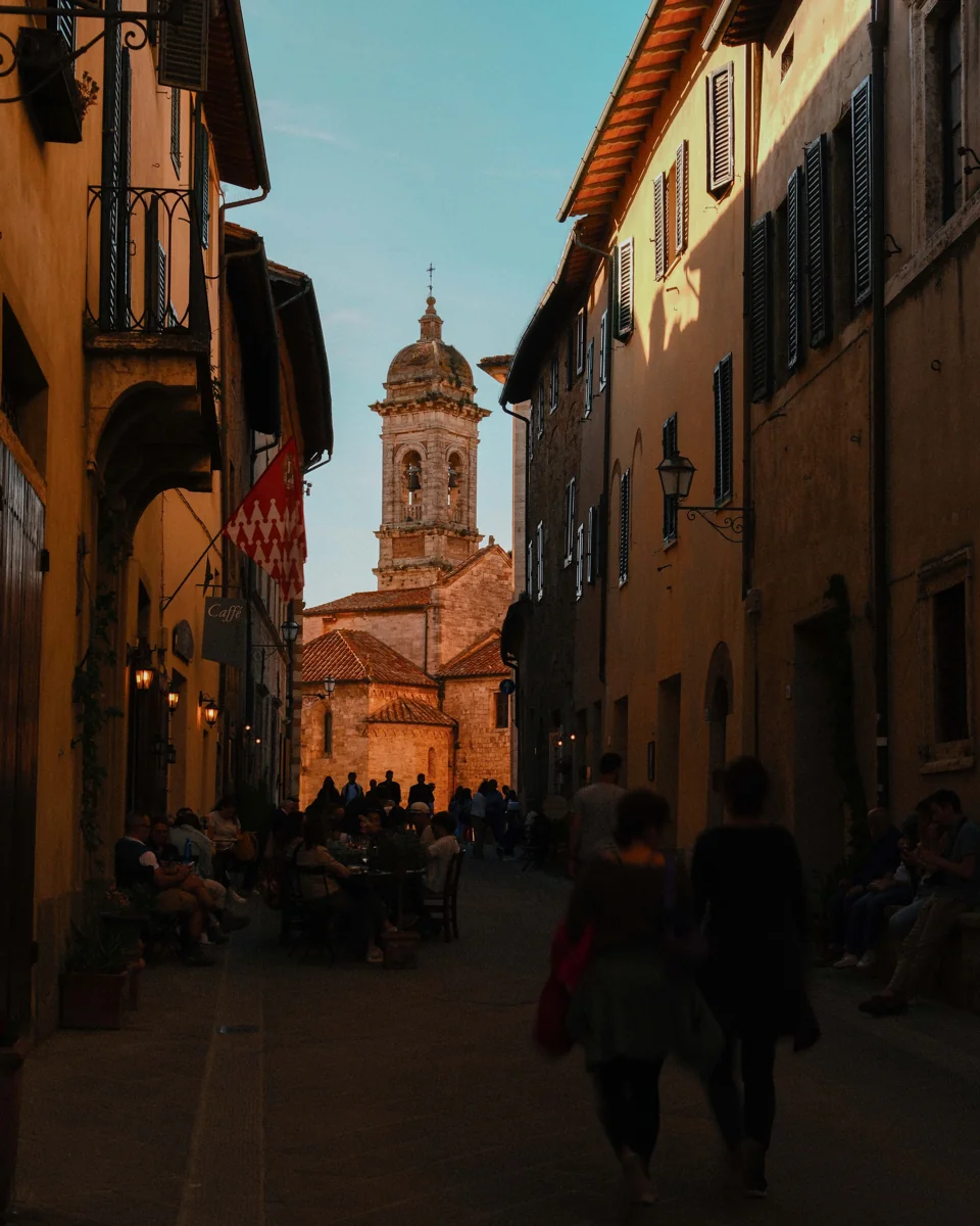 A shaded street opening toward a sunlit church tower, with people walking in the foreground.