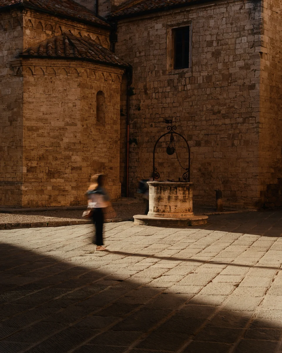 A person crossing a stone courtyard beside a well, through a band of sunlight.