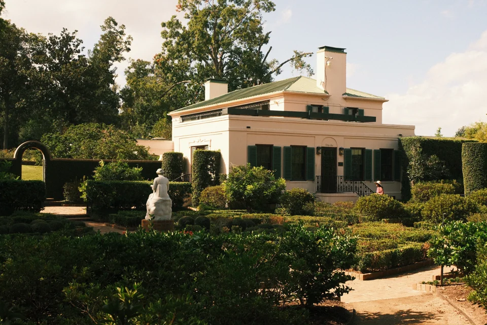The pale house with dark green shutters beyond the patterned hedges of the garden.