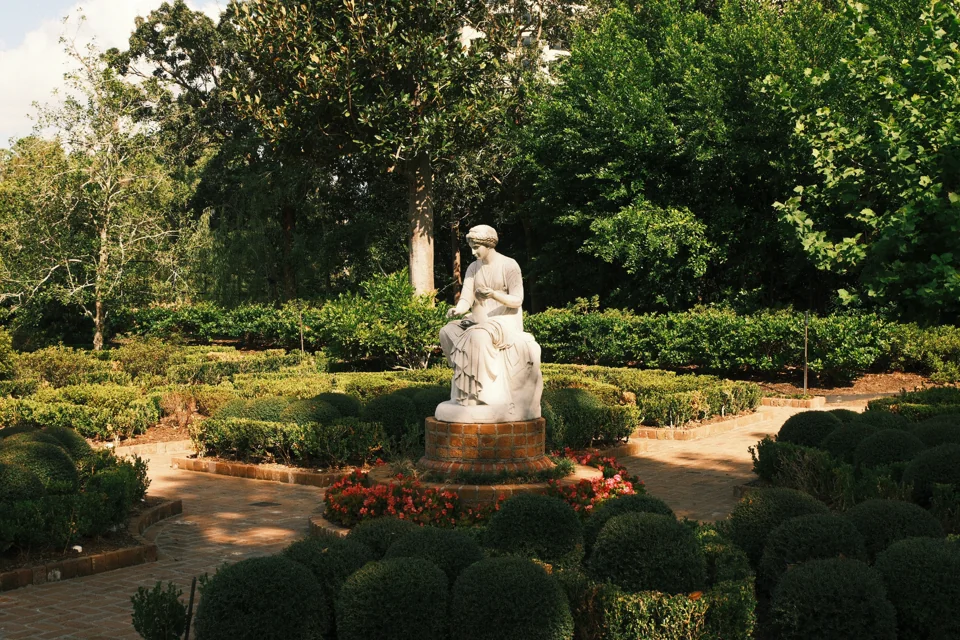 A white seated sculpture at the meeting of garden paths, surrounded by rounded hedges and trees.