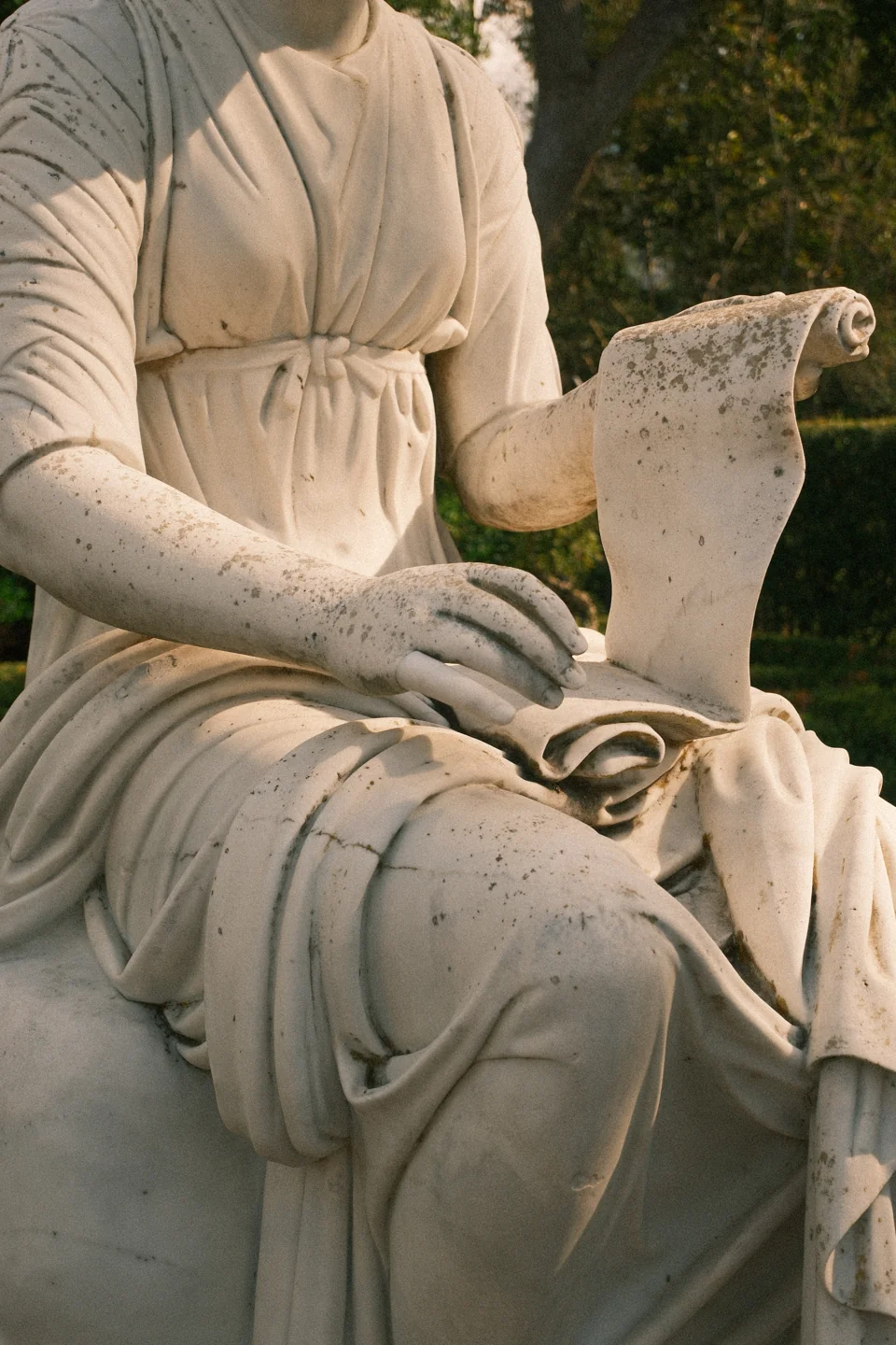 A close view of a seated sculpture’s hand, scroll, and softly folded stone drapery.