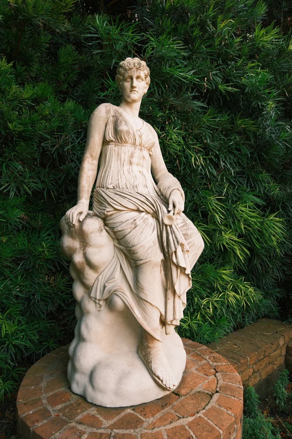 A standing stone figure in draped clothing on a round brick base among dense greenery.