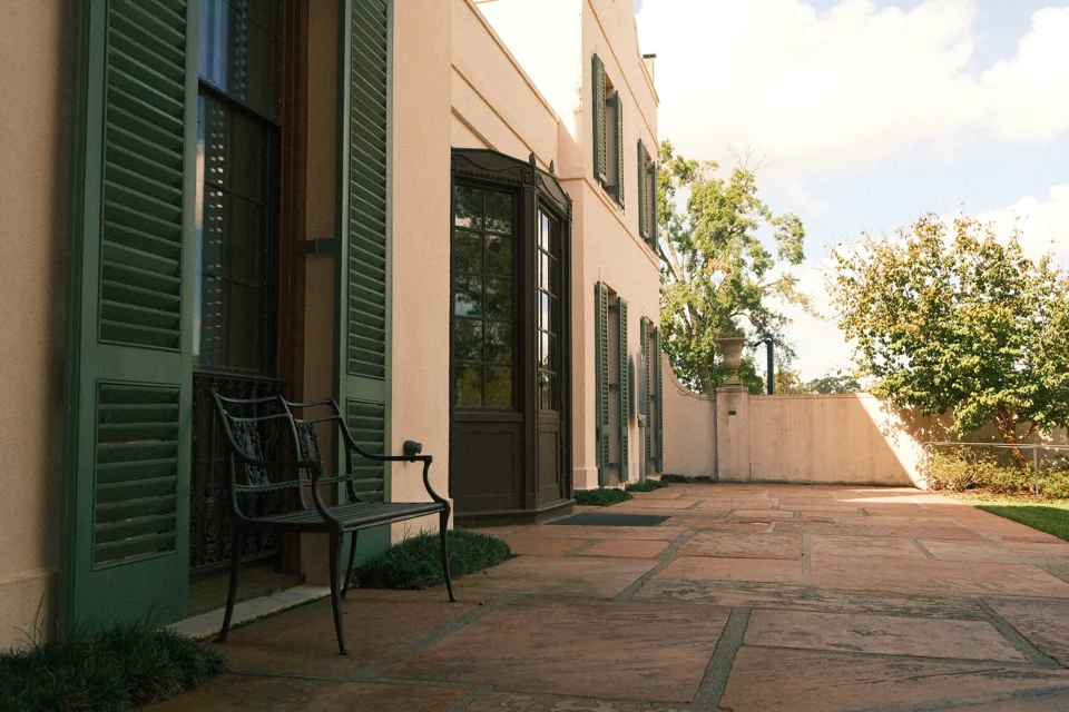 An empty chair beside tall green shutters on the sunlit brick terrace.