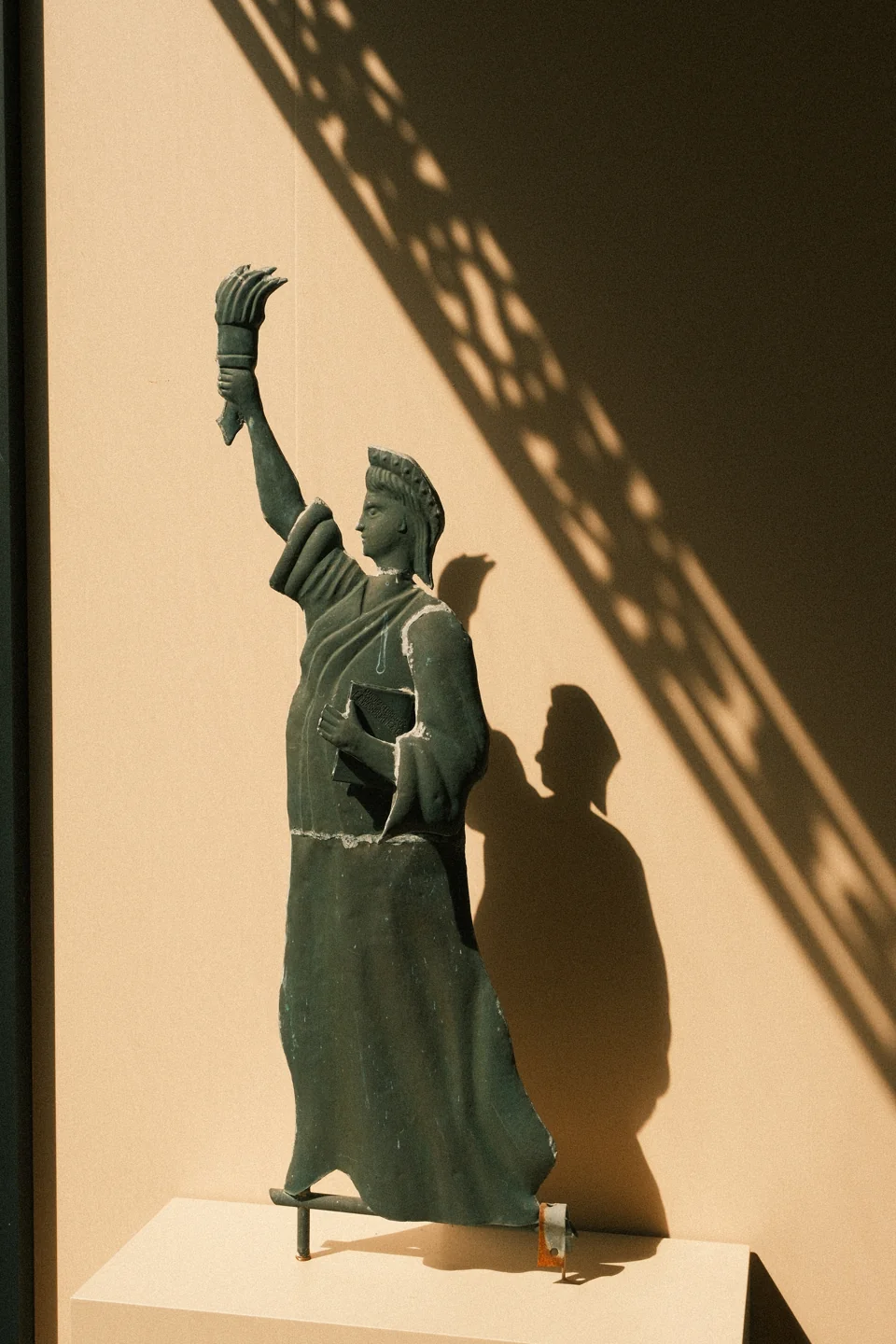 A small raised-arm sculpture beside a patterned shadow on a sunlit interior wall.